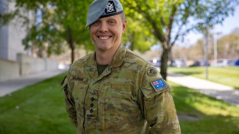 A man in uniform with a blue beret smiles with greenery and buildings behind him.