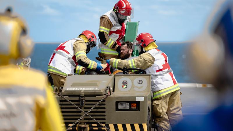 Three people in firefighter uniforms and red helmets wearing Red Cross vests over the uniforms attend to a man seated in an open-roofed vehicle on the deck of a ship.