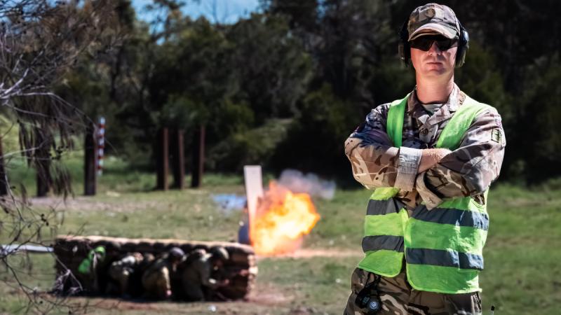 A uniformed soldier wearing a high-visibility vest and sunglasses stands with arms crossed as a fireball rises behind sandbags.