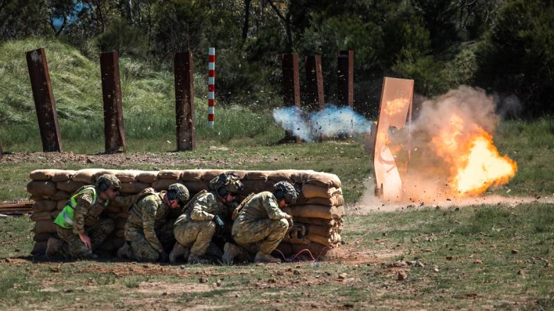 Soldiers crouch behind sandbags as a small fireball rises from a detonation nearby.
