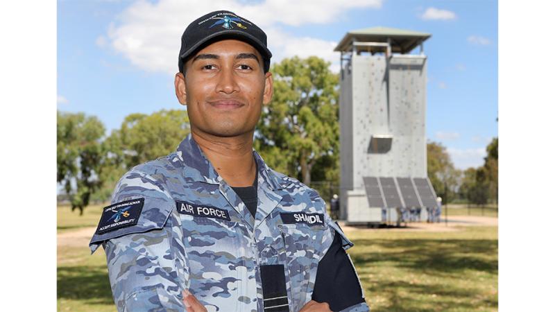 A man in Air Force camouflage uniform and cap standing outdoors, a rock climbing wall in the background.