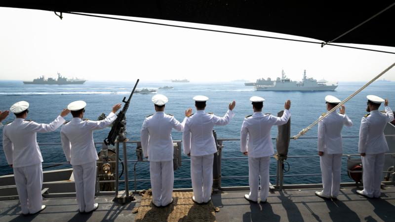 People in uniform standing on the deck of a ship wave to nearby ships and boats.
