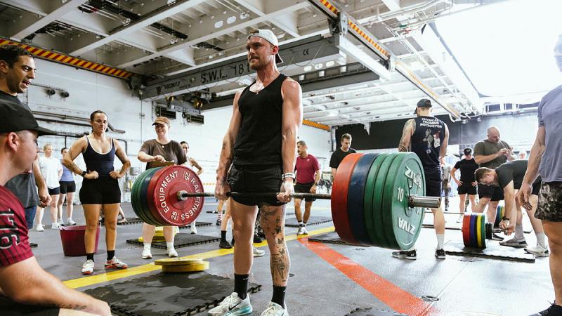 A Navy member stands holding a barbell full of weights with others watching on.