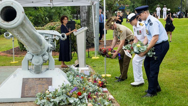 Three Defence personnel, one each from Navy, Army and Air Force hold wreaths to lay at a cannon-style weapon memorial.