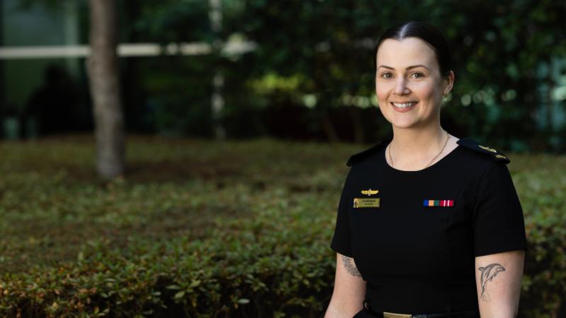 A woman in Navy uniform outside near a hedge, with a large building in the background.