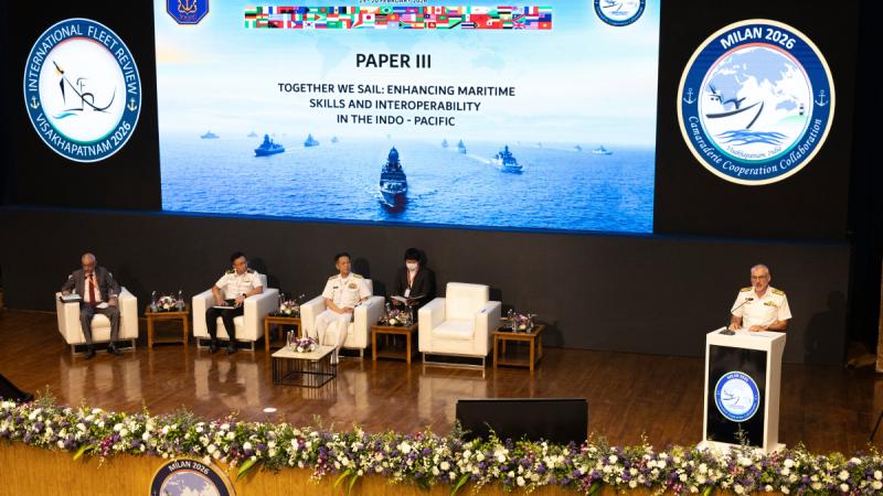 A man in uniform stands behind a podium on a stage with four people seated nearby and a large screen above them.