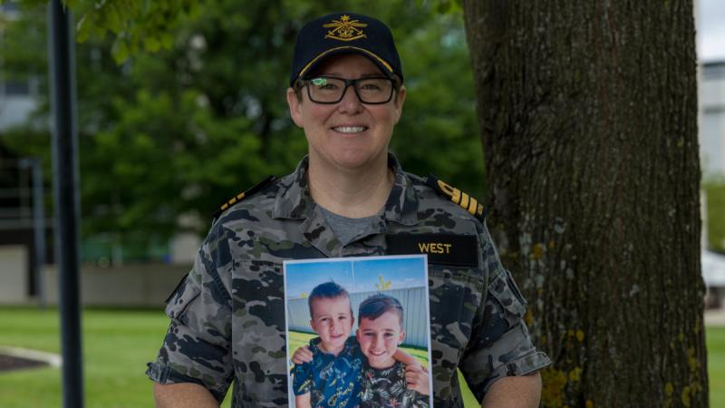 A woman in Navy uniform stands outdoors holding a printed photo of two boys.