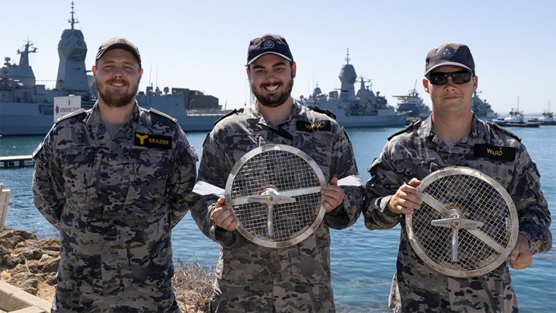 Three military men hold two metal strainer devices by the ocean.