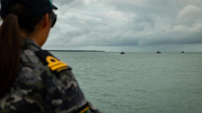 A uniformed woman seen from behind watches three ships sailing out of a harbour.