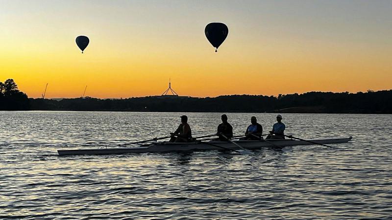 Four rowers sit in their rowboat under a golden sunrise with two hot air balloons in the sky behind them.