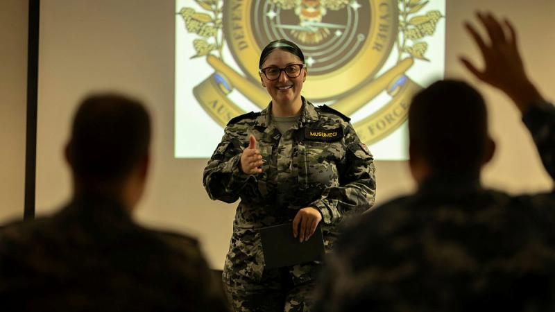 A Navy member stands in front of an emblem facing others. One person in the room has their hand raised.