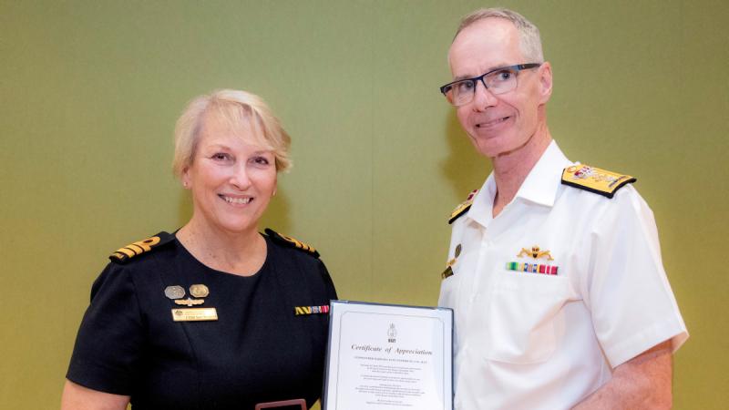 A woman in uniform holds a presentation box while standing beside a man in uniform holding a framed certificate.