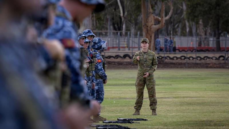 A woman in green camouflage stands facing a line of people in blue camouflage uniforms with rifles resting on the ground.