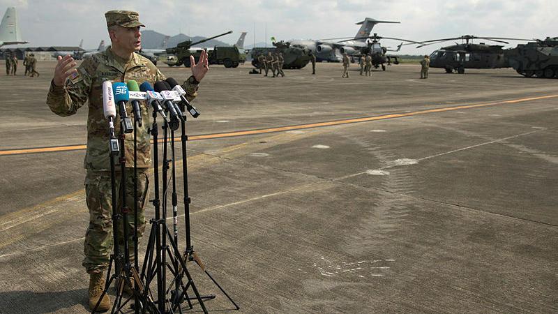 A man in uniform stands outdoors in front of microphones.