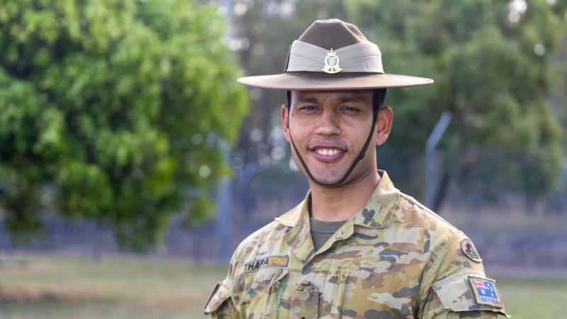 A man in Australian Army camouflage and a broad brim uniform hat stands outdoors.