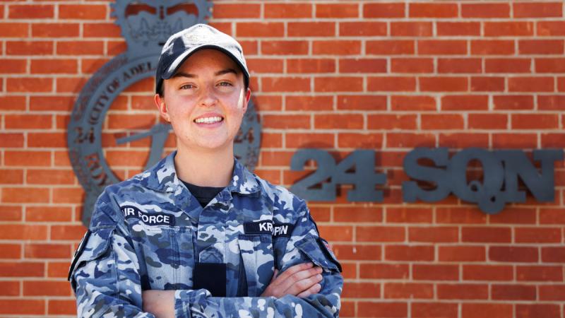 A woman in Air Force uniform stands outside in front of a brick wall with a metal emblem and the letters 24 SQN on it.