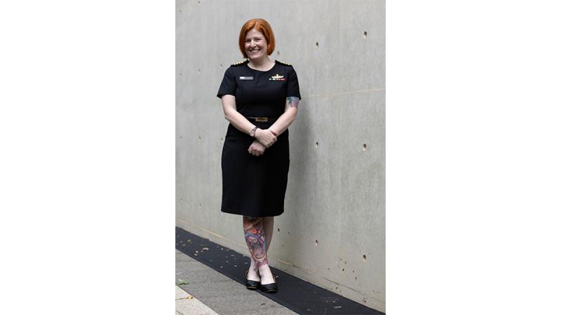 A woman in Navy uniform stands in front of a concrete wall outdoors.