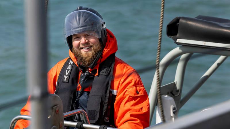 A Navy member in an orange cold-weather jacket and head protection smiles, with ocean in the background.