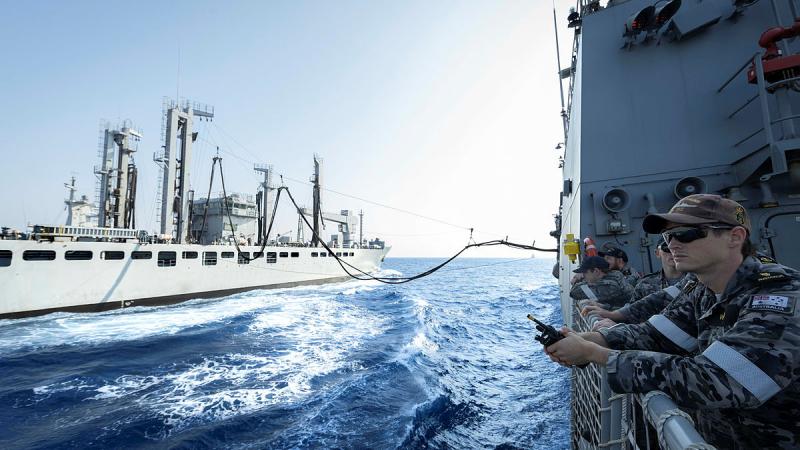 Two ships are joined by a fuel line. Navy personnel on one of the ships look out to sea.