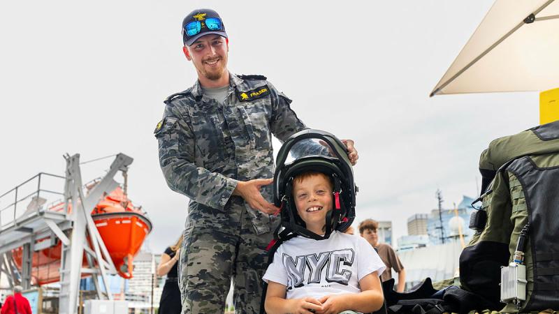 A Navy member places clearance diving headwear onto a young smiling child who is sitting on a chair.