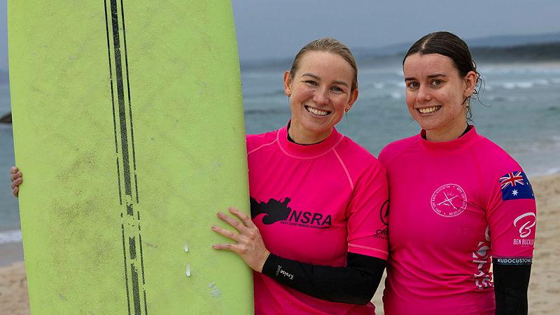 Two women in pink rash vests stand next to a bright yellow surfboard on a beach.