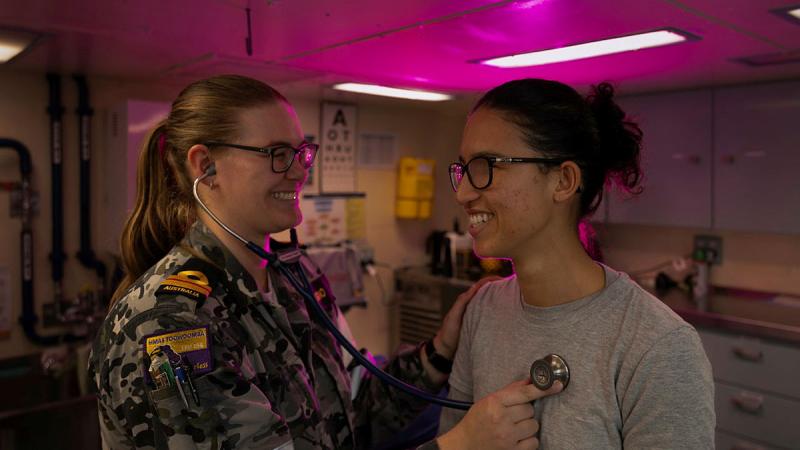 A Navy medic places a stethoscope on the chest of a patient in a pink-lit room.