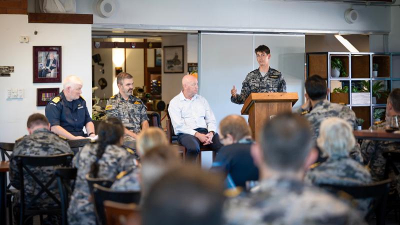 A man in camouflage uniform stands at a lectern, gesturing to three men sitting beside him in a medium-sized room facing an audience.