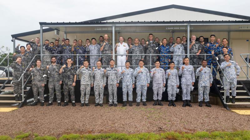 A group of people in uniform stand in three rows in front of a large demountable building, their right fists raised.