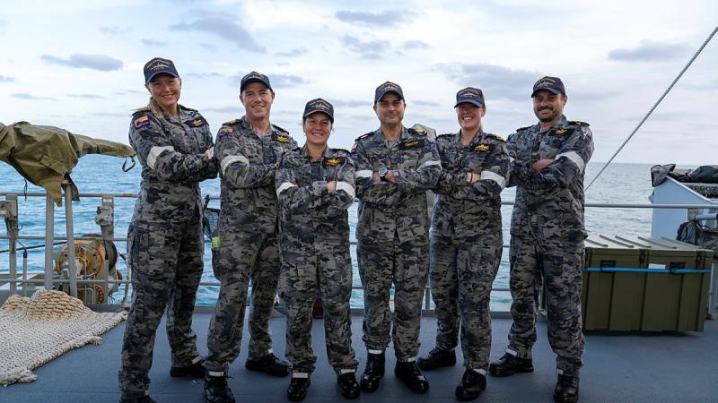 Six Navy personnel in grey/black uniforms stand on the deck of a ship with their arms crossed.