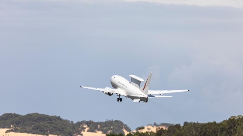 An aircraft climbs through the sky above trees.