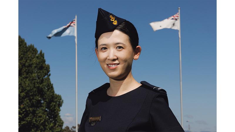 A woman in military uniform stands outside with two national flags.