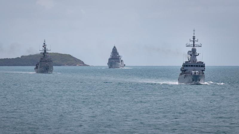 Four naval ships sail in close formation at sea, with a low green peninsula visible in the background.