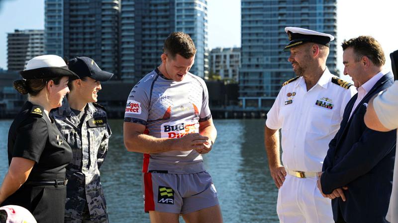 A group of people, including a rugby league player and Navy personnel stand by a body of water with buildings in the background.