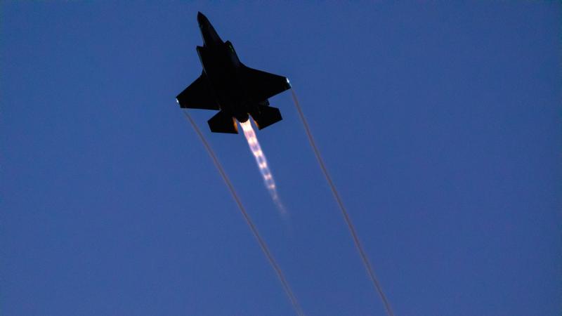 A jet aircraft flies through a clear blue sky with visible exhaust flame and vapour trails.