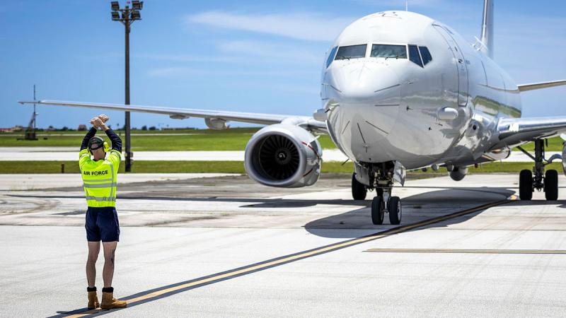 A person stands in yellow high-vis gear with arms up and crossed, signalling an aircraft.