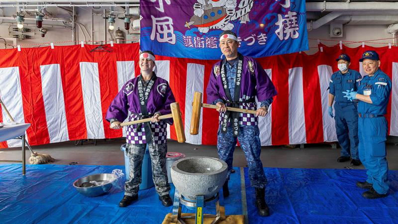 Two people stand in purple outfits, holding mallets in front of a red and white striped background.