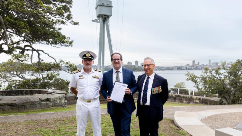 Three men stand in front of a naval memorial, with one holding a document, and Sydney Harbour in the background.
