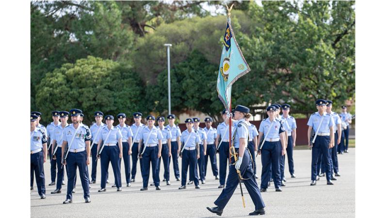 A military woman holds a flag aloft as she walks past military members standing at attention. 