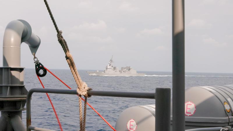 A ship sails ahead in open water, viewed from another ship’s deck with railings and equipment visible.