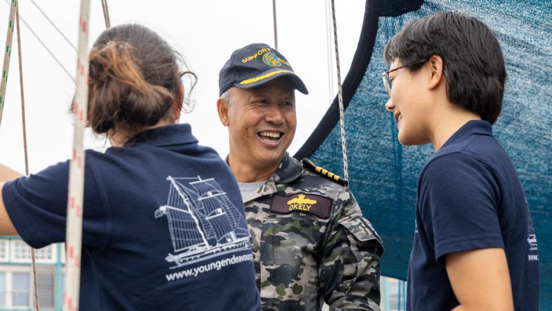 A man in Navy uniform speaks to two young women in blue polo shirts, with several ropes dangling around them and a blue shade sail behind him.