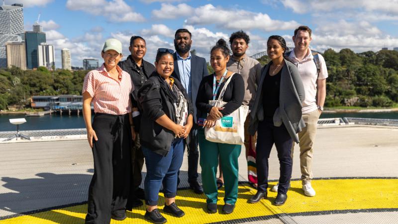 A group of people standing on a ship’s helipad with Sydney Harbour Bridge and the city skyline in the background.