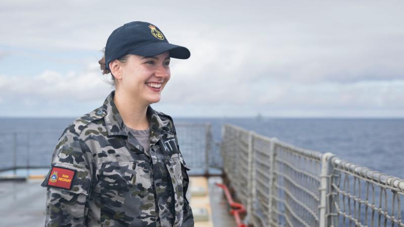 A woman in a camouflage Navy uniform, with a red patch reading ‘RAN Recruit’ on her shoulder, stands on the deck of a ship smiling out to sea.