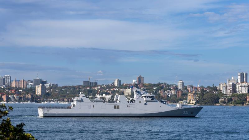 A ship sailing in a harbour, buildings and greenery on the shore in the background.
