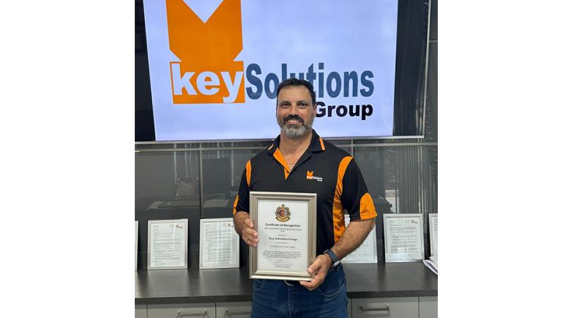 A man in a polo shirt and jeans stands indoors holding a framed certificate.