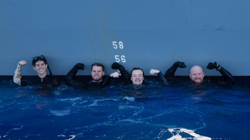 Four divers make strong arms above their heads near the side of a ship and they tread water.