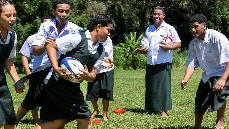 A Tongan student in uniform runs with the ball as others look on and give chase.