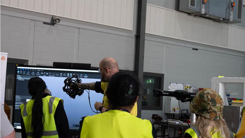 Students in high visibility vests watch a person demonstrate a handheld device connected by cables to a large screen in a large room.