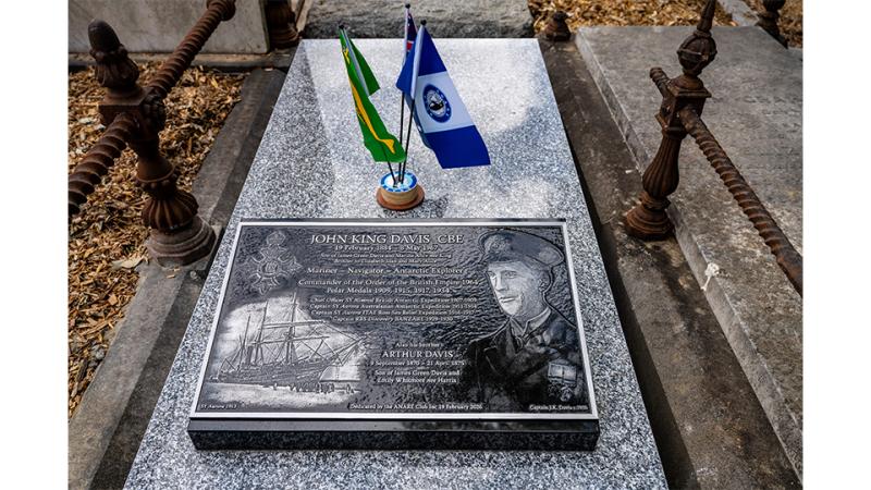 Grey stone headstone with a detailed plaque featuring a carved ship, a crest and a portrait, with three small flags placed above it.