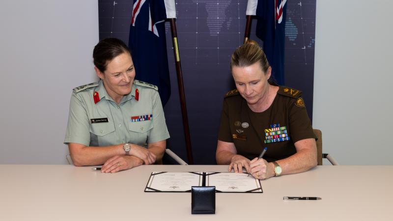 Two women in military uniform sign a document with New Zealand and Australian flags behind them.