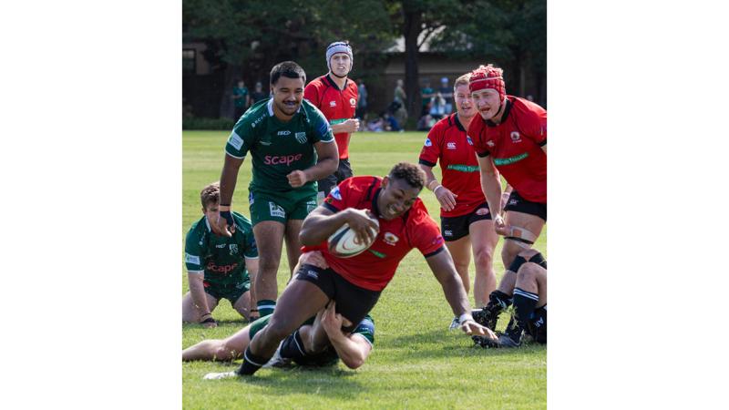 A group of men play rugby, with one man holding the ball getting tackled.